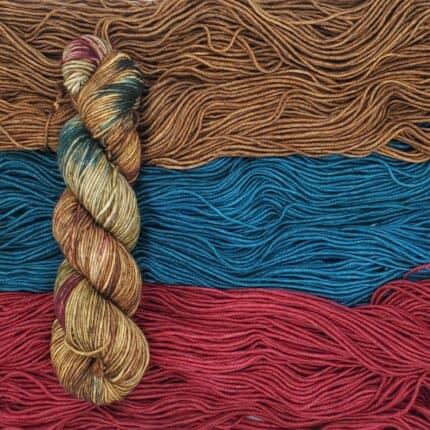 An uneven earth tone variegated skein of yarn atop semisolid skeins of a rich medium brown, vibrant blue, and deep red.
