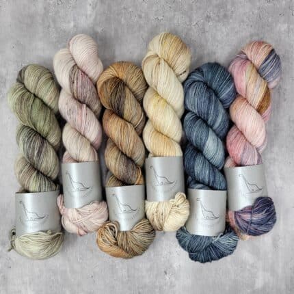 Six skeins of yarn in a khaki green, cream speckled with orange and pink, a dusty yellow, orange and brown, cream layered with orange and brown, a teal blue variegated and a soft pink layered with blue and tan.