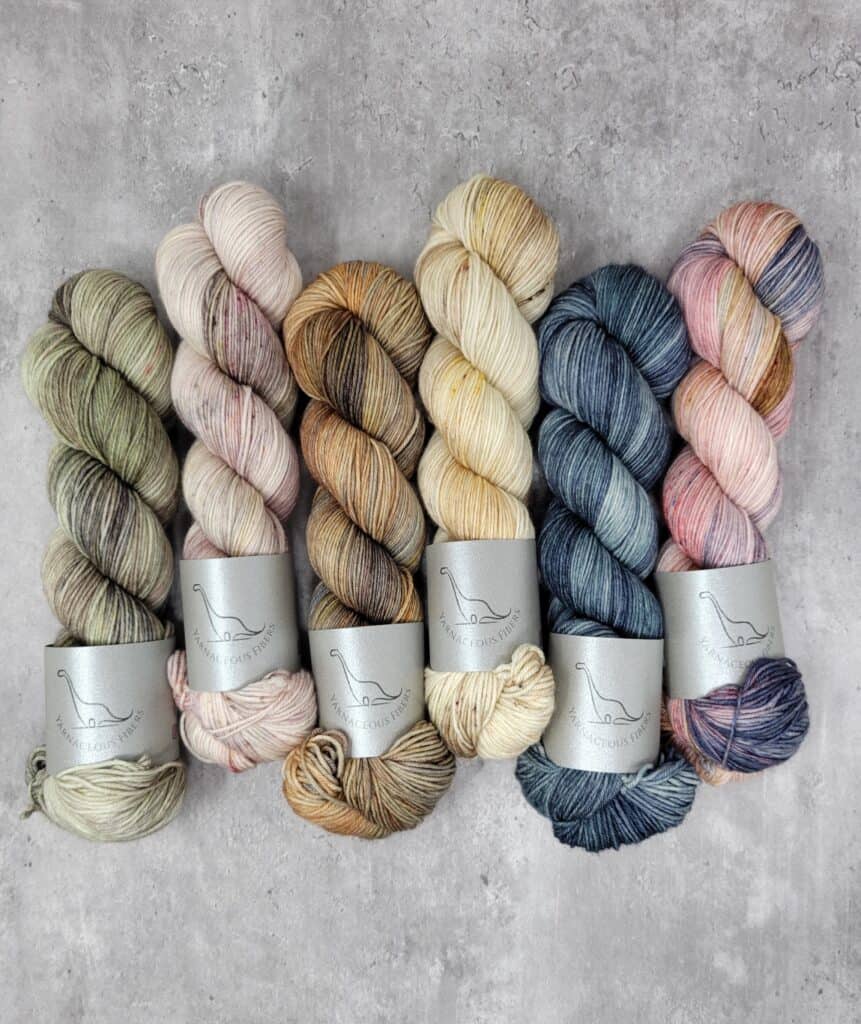 Six skeins of yarn in a khaki green, cream speckled with orange and pink, a dusty yellow, orange and brown, cream layered with orange and brown, a teal blue variegated and a soft pink layered with blue and tan.