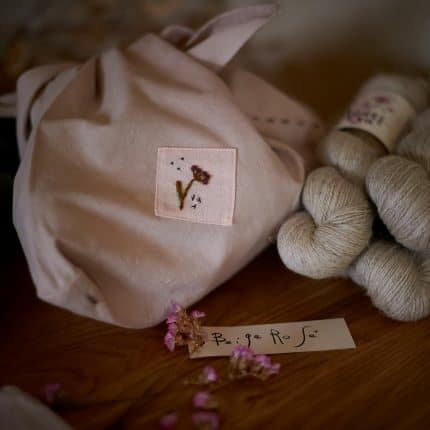 A pale pink hand-dyed and embroidered knitting bag with dried flowers and un-dyed yarn around it.