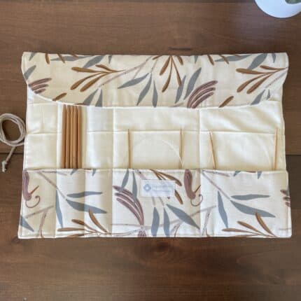 A partially open knitting needle roll with exterior leaves print and a cream colored lining visible. There are various wooden needles in the slots.