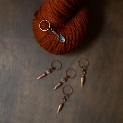 A stitch marker with a shovel charm sits on a hank of burnt orange yarn. Four carrot stitch markers are scattered on the table below.