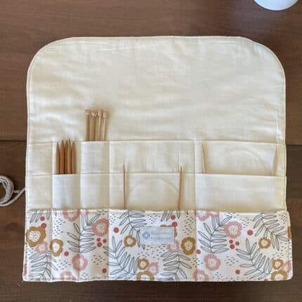 A fully open knitting needle roll with exterior floral print and a cream colored lining visible. There are various wooden needles in the slots.