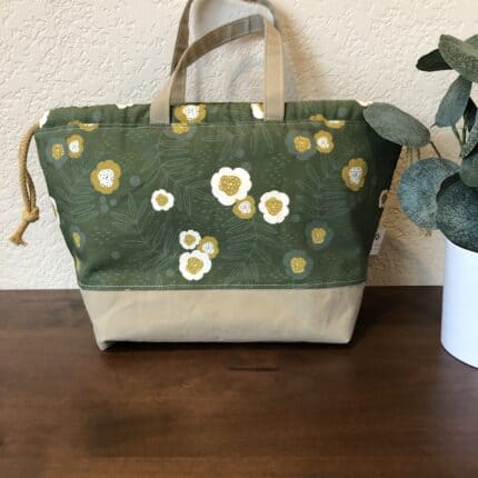 A canvas drawstring bag with cream flowers on a green background. There is a beige bottom with matching beige handles.