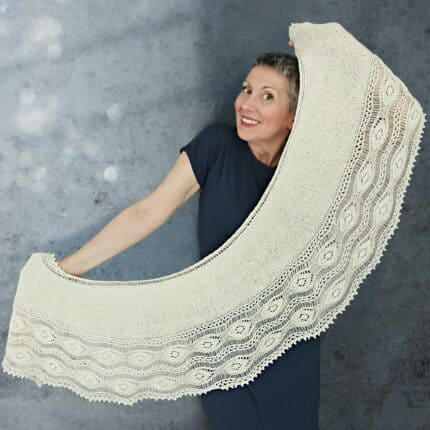 A white woman holding a shawl with both hands. The shawl has a half donut shape and a lacy border.