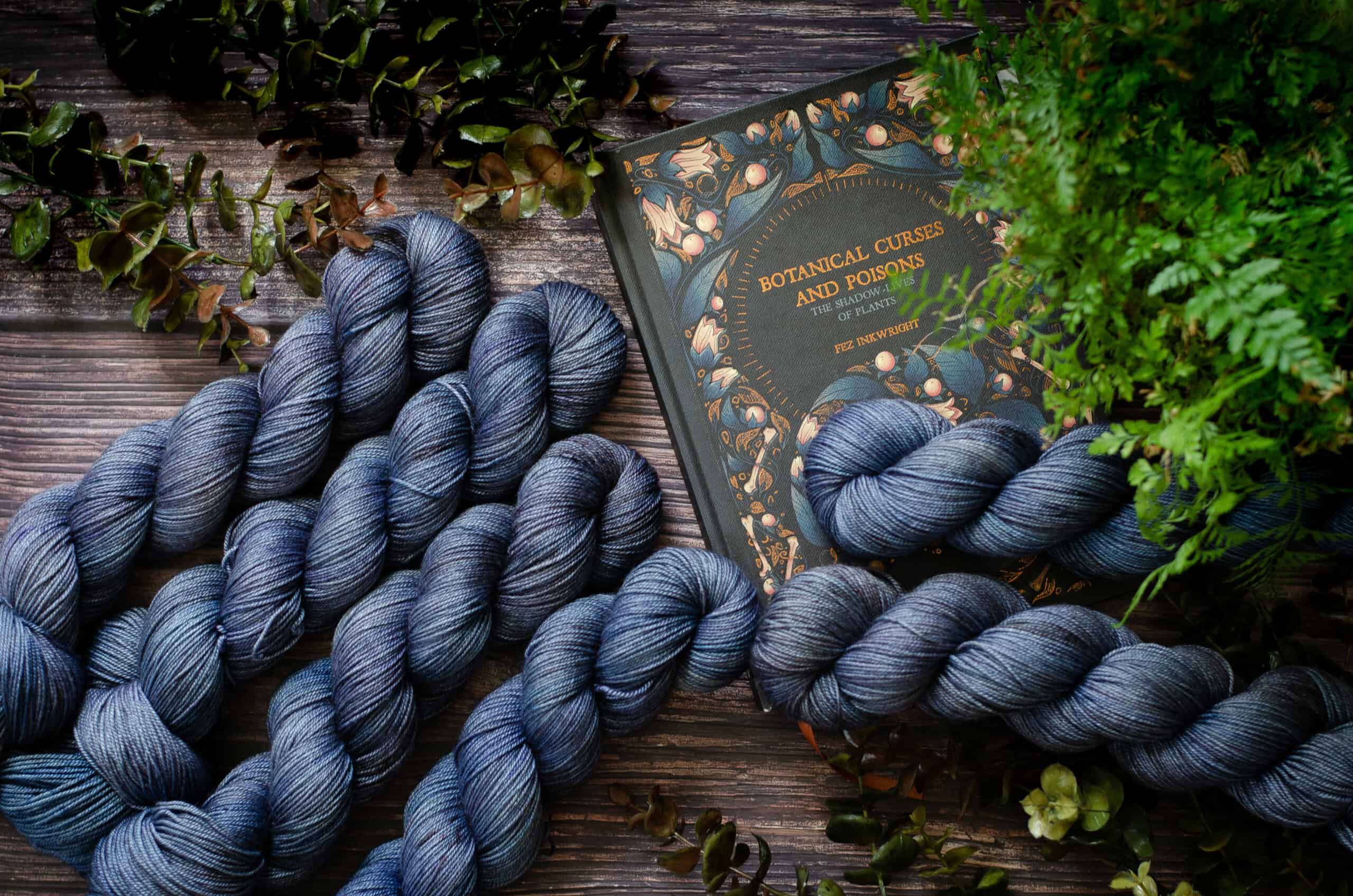 Six skeins of blue-purple yarn surrounded by ferns and a book with the title Botanical curses and poisons.