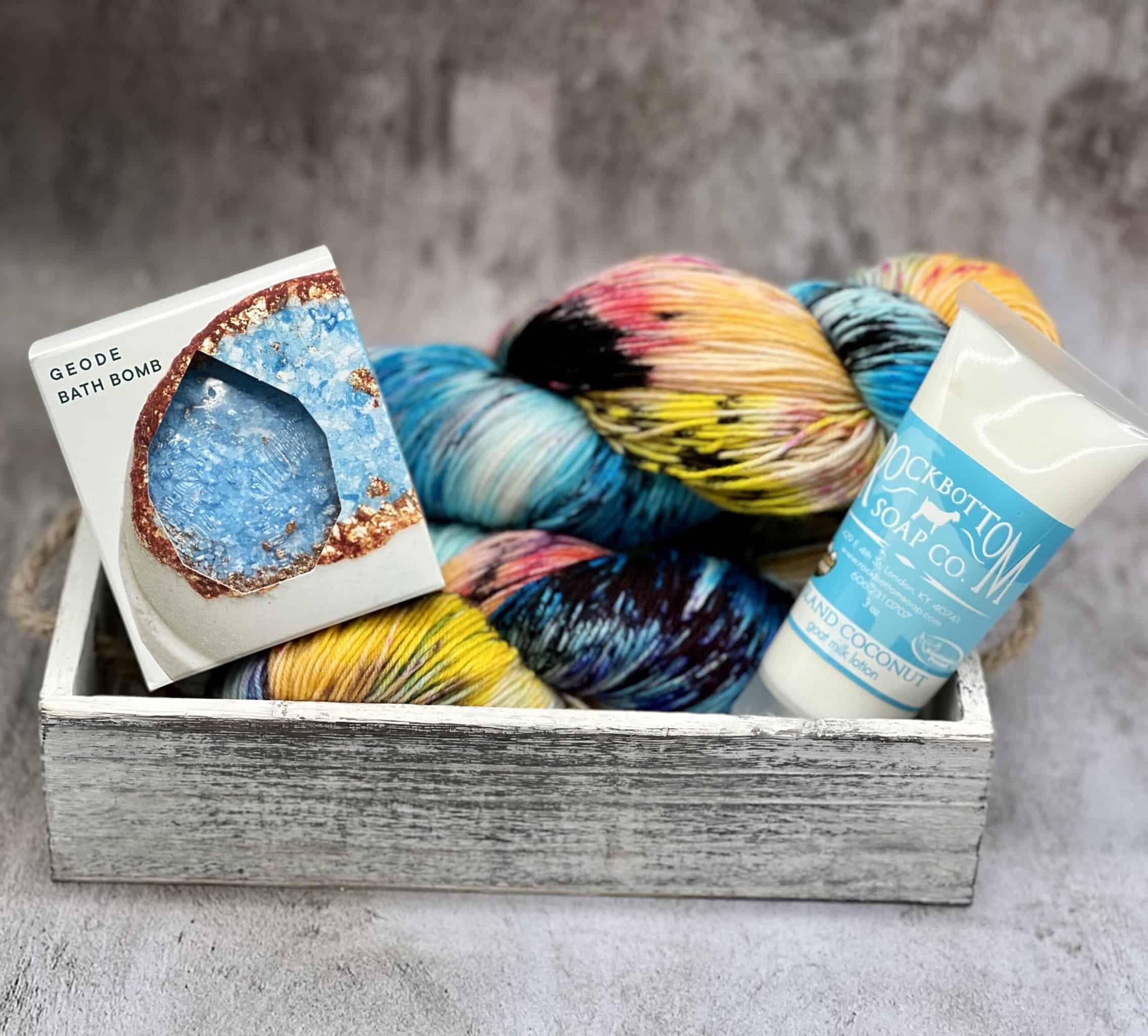 A skein of yarn in blue, yellow, red and black with a blue bath bomb and coconut lotion in a white wooden box.