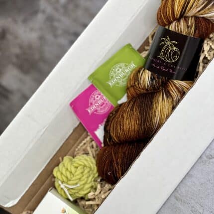 A skein of yarn in shades of brown in a white cardboard box, with two tea bags, a light green yarn candle and a lip scrub.