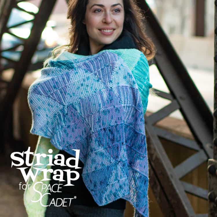An oblong wrap knit in rows of modular triangles that change in a gradual gradient along the length of the piece, and the words Striad Wrap SpaceCadet.