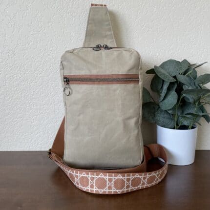 A beige canvas sling backpack with a caramel colored strap in a geometric pattern.
