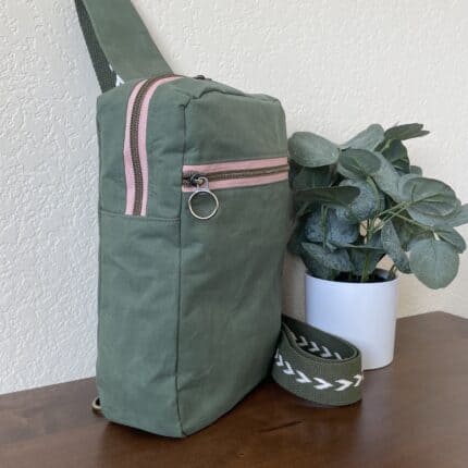 A olive green canvas sling backpack with a dusty pink zipper.