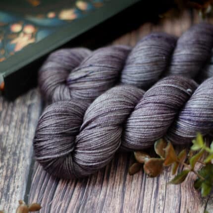 Two skeins of lilac yarn surrounded by plants on a wood surface, a dark blue book is in the background.