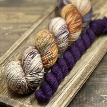 A skein of variegated neutral colored yarn with a deep purple mini.