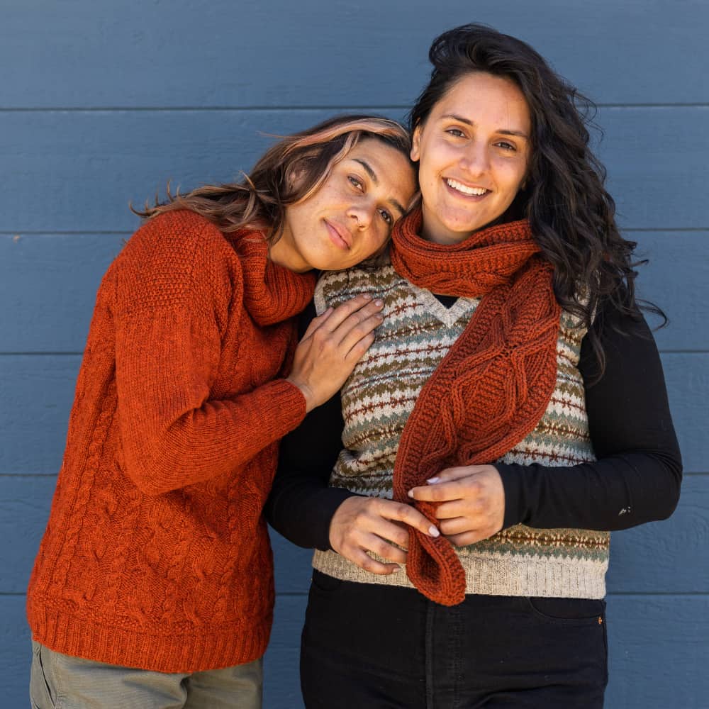 Two women wearing a red textured sweater, fairisle colorwork vest and cabled scarf in rusty reds and earthy neutrals in front of a blue wall.