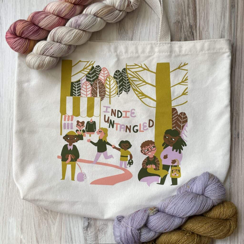 A tote bag with a lilac, pink, gold and gray illustration of a diverse group enjoying a fiber festival.