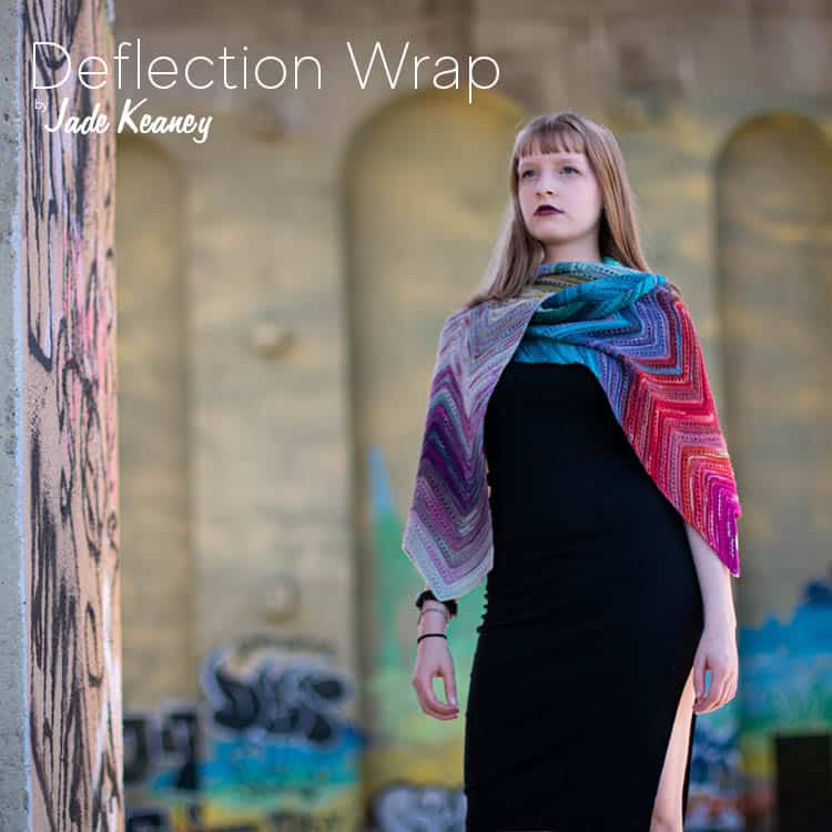 A young light-skinned woman in a black dress and a colourful chevron wrap, standing underneath a graffiti'd bridge and gazing upward with a look of strength on her face, and the text Deflection Wrap by Jade Keaney.