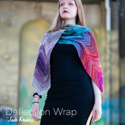 A young light-skinned woman in a black dress and a colourful chevron wrap, standing underneath a graffiti'd bridge and gazing upward with a look of strength on her face. The text Deflection Wrap by Jade Keaney.