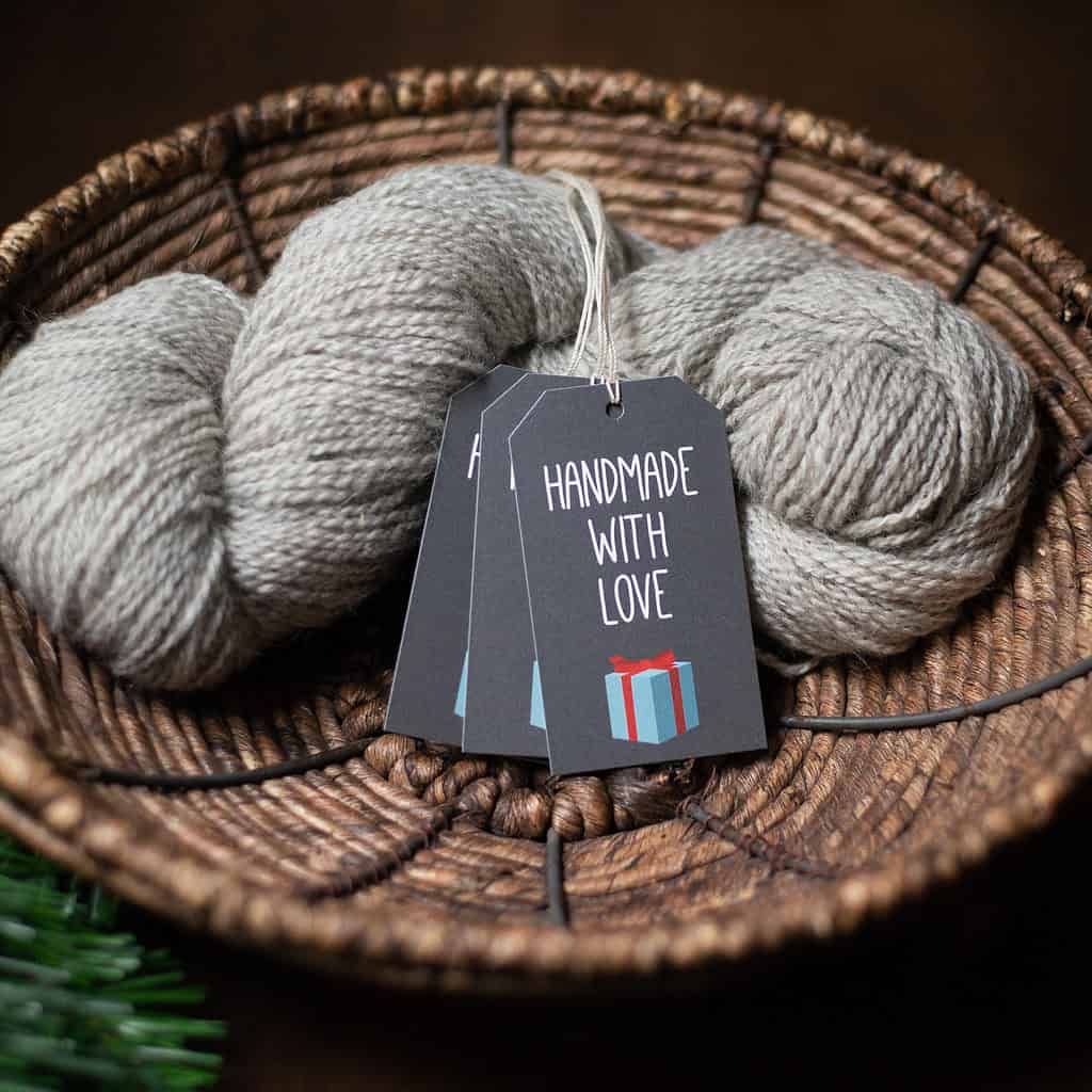 Dark green gift tags that say "handmade with love" rest on a skein of light gray yarn sitting in a woven basket.