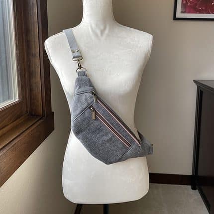 A grey flannel crossbody sling bag with pink zips is pictured hanging on a mannequin.