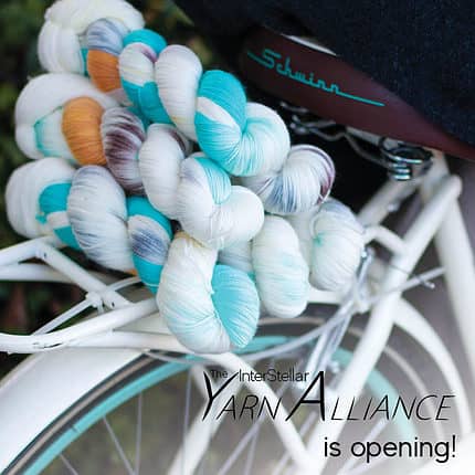 A pile of fingering yarn on the luggage rack of a bike, in a variegated colourway of teal, honey, brown, grey, and natural, and the text The Yarn Alliance is opening.