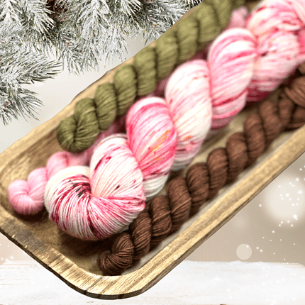 A large skein of red, white, and brown yarn with three mini skeins in pink, brown and green under Christmas decor.