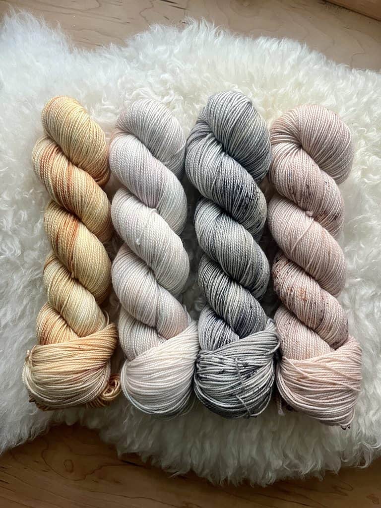 Four skeins of softly variegated yarn, golden yarn, pastel yarn, gray and pale yellow with speckles, and a tan tonal with multicolor speckles.