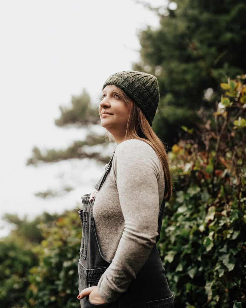 A light-skinned woman in a green knit cap stands near bushes.