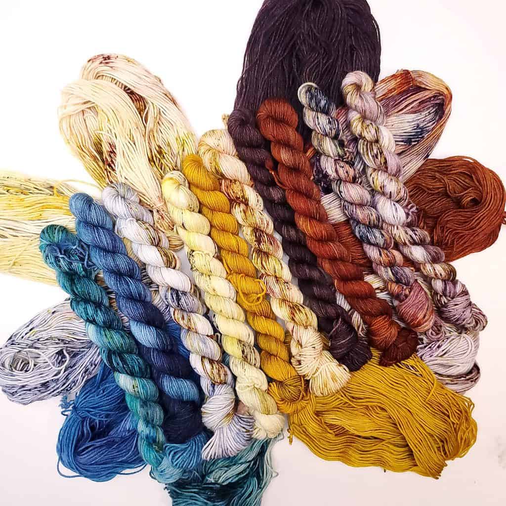 Mini skeins in blue, gold, red and purple arranged in a row resting on top of loose hanks of yarn of the same colors.