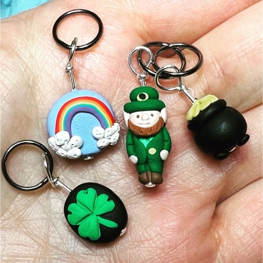 Four St. Patrick’s Day themed polymer clay stitch markers. The set includes a rainbow on a blue background, a bright green four leaf clover on a black background, a leprechaun dressed in green, and a pot of gold coins.