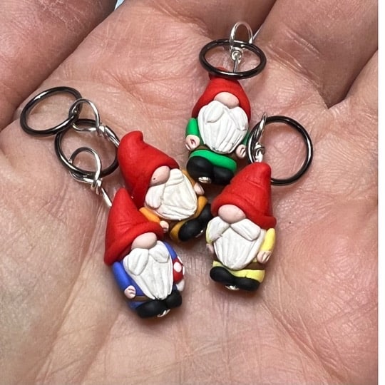 Four hand-sculpted garden gnome stitch markers wearing hats that are so large they’ve slipped over their eyes. Each gnome is wearing a different colored shirt - orange, blue, yellow, and green. One gnome is carrying a small red and white mushroom.