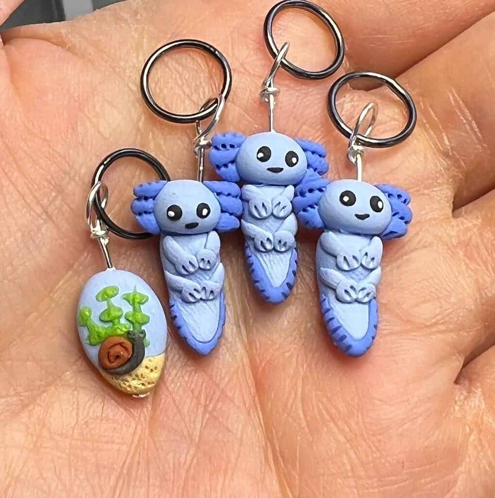 Three miniature blue axolotyl stitch markers and one stitch marker featuring an aquatic plant with a tiny snail.