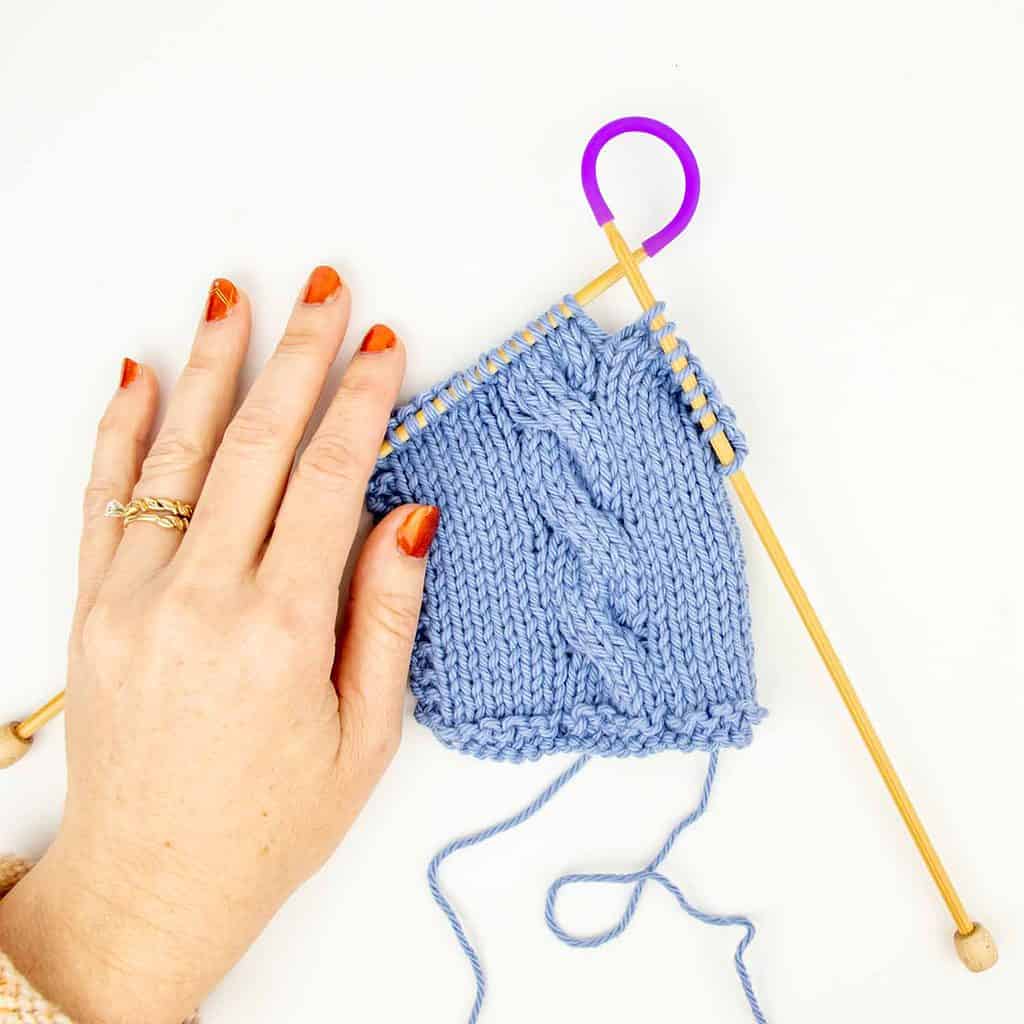 A light-skinned hand with blue yarn on brown knitting needles with purple tip ties.