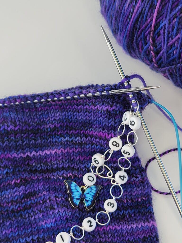 Blue butterfly chain row counter on purple knitting with a white background.