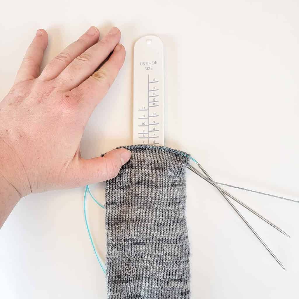 Grey knitted sock with a white sock ruler on a white background with a light-skinned hand.
