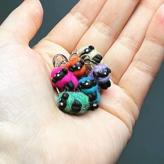 Six Woolly Sheep Stitch Markers sit in an open, light-skinned hand. Their fleeces are the color of the rainbow.