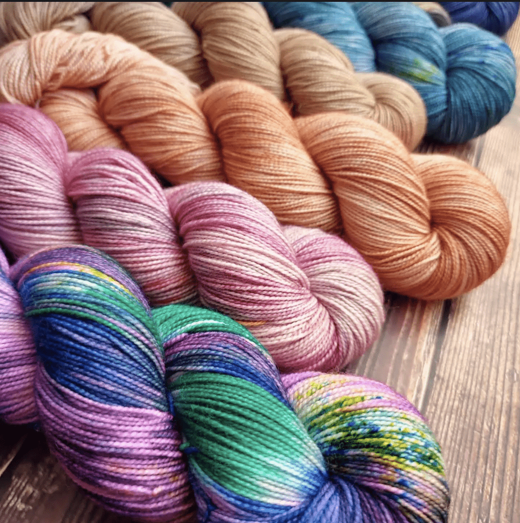 Skeins of blue, blush, light pink, and variegated blue, purple, and green skeins laying on a wooden table.