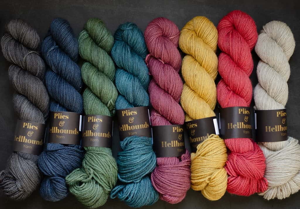 A line up of eight skeins, charcoal navy, deep green, teal, wine red, golden yellow, poppy red and a natural tinted cream.