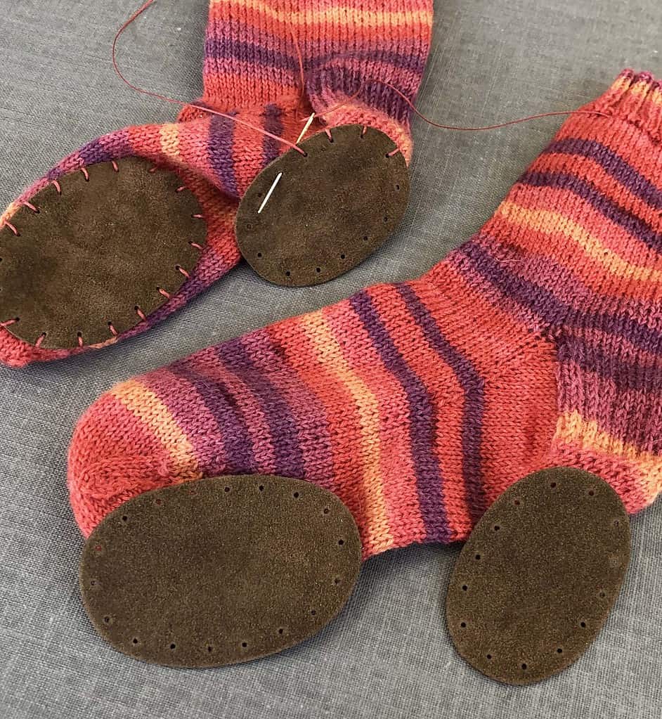 A handknit striped sock in pink and purple with brown suede leather patches stitched on.