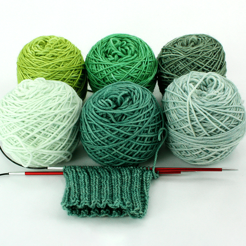 Cakes of yarn in various shades of green and green ribbing on red needles.