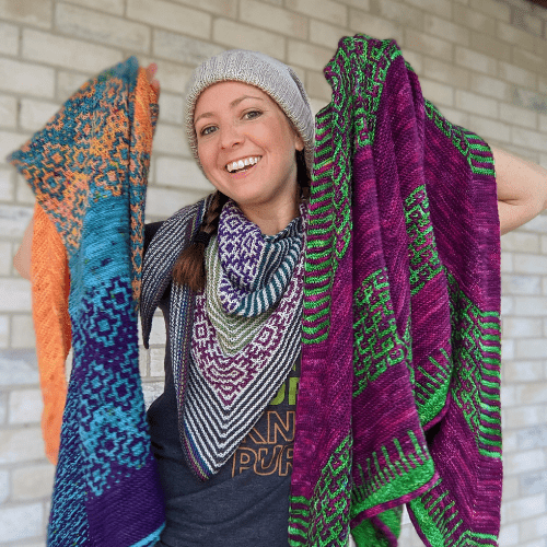 A light-skinned woman wearing a grey beanie and holding several colorful shawls.