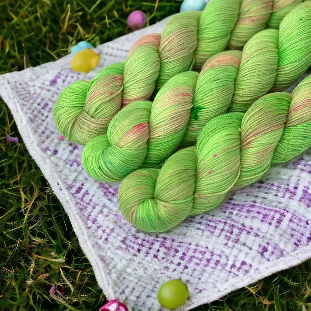 Three skeins of bright green yarn with pink speckles laying on a while and purple cloth on grass.
