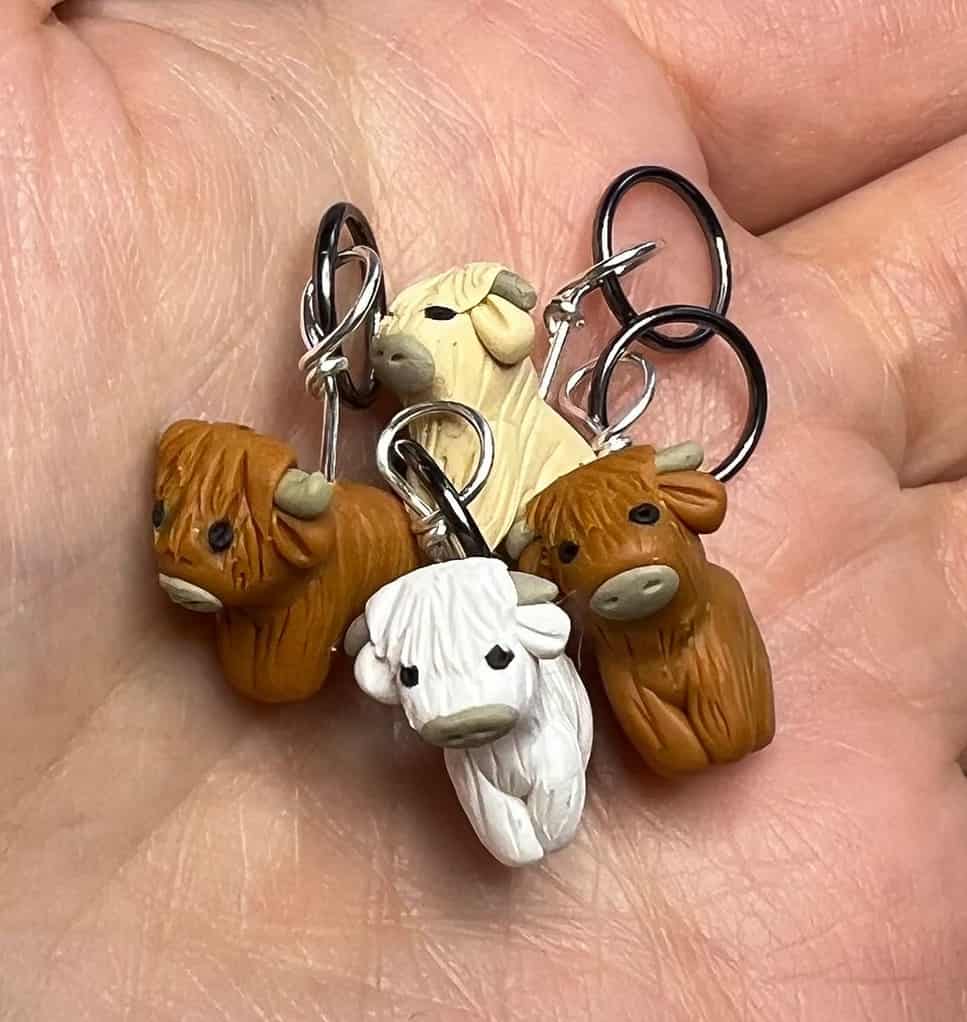 Four Highland coo stitch markers. One cow is white, one tan, and two are a deeper brown with hints of orange.