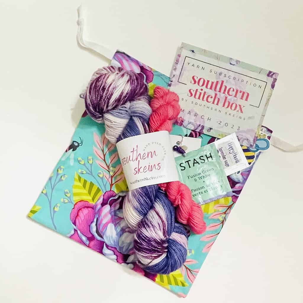 A skein if blue, purple and white hand-painted yarn packaged with a pink mini skein, on top of a bright floral bag and a card that reads Southern Stitch Box.