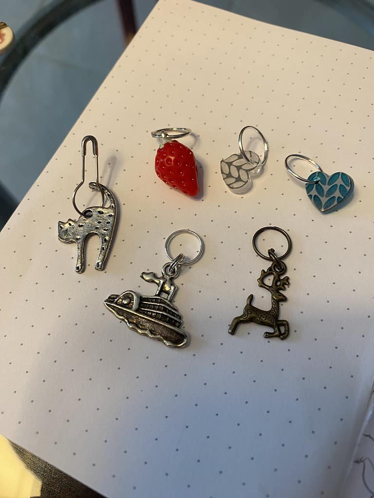 Set of 6 stitch markers: a silver cat, boat and stag, a strawberry and clear and blue hearts with stockinette stitch.
