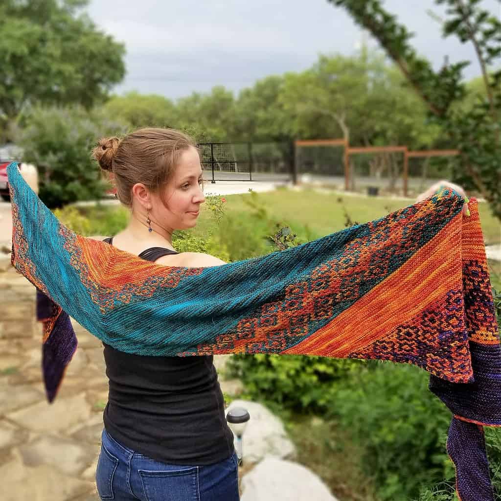 A light-skinned woman standing outdoors holding a long teal, orange and purple shawl behind her back.
