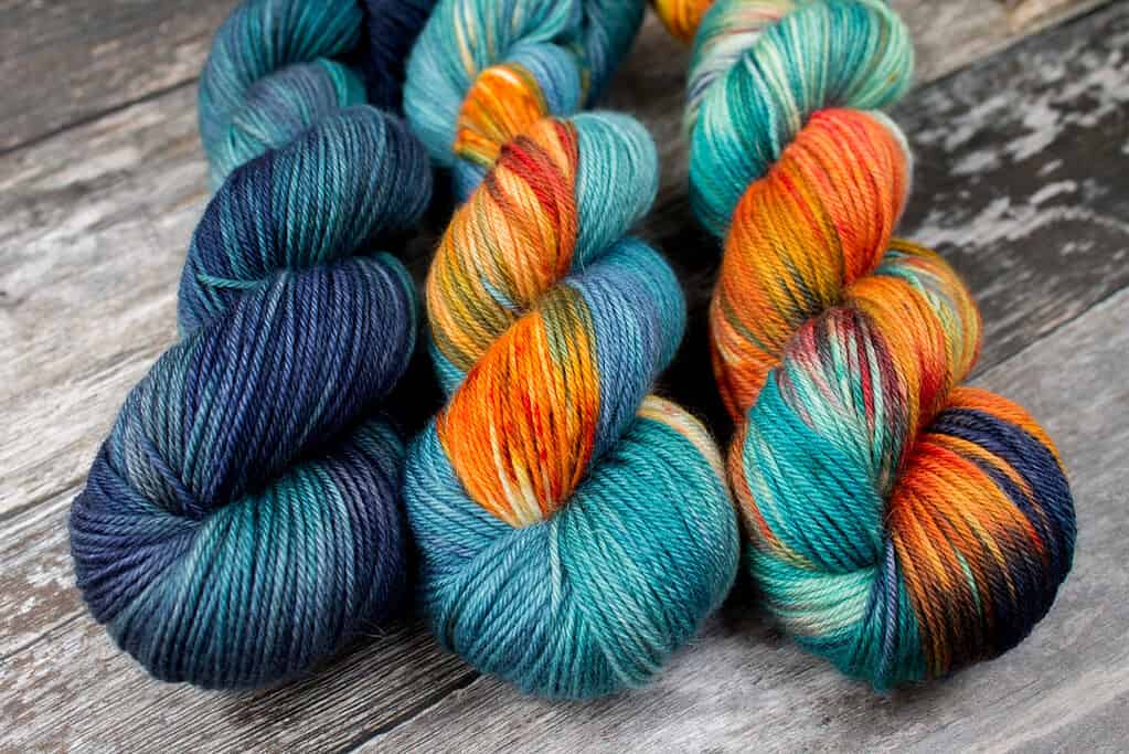 Three skeins, in navy and teal, light navy teal with orange speckled sections, and variegated teal navy and oranges with gold.