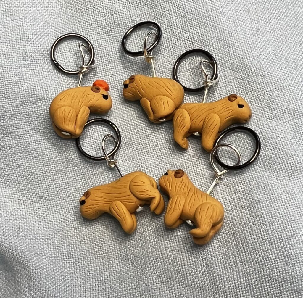 Five capybara stitch markers. Two are standing, three are sitting. One of the sitting capybaras is balancing a tiny orange carefully on his head.