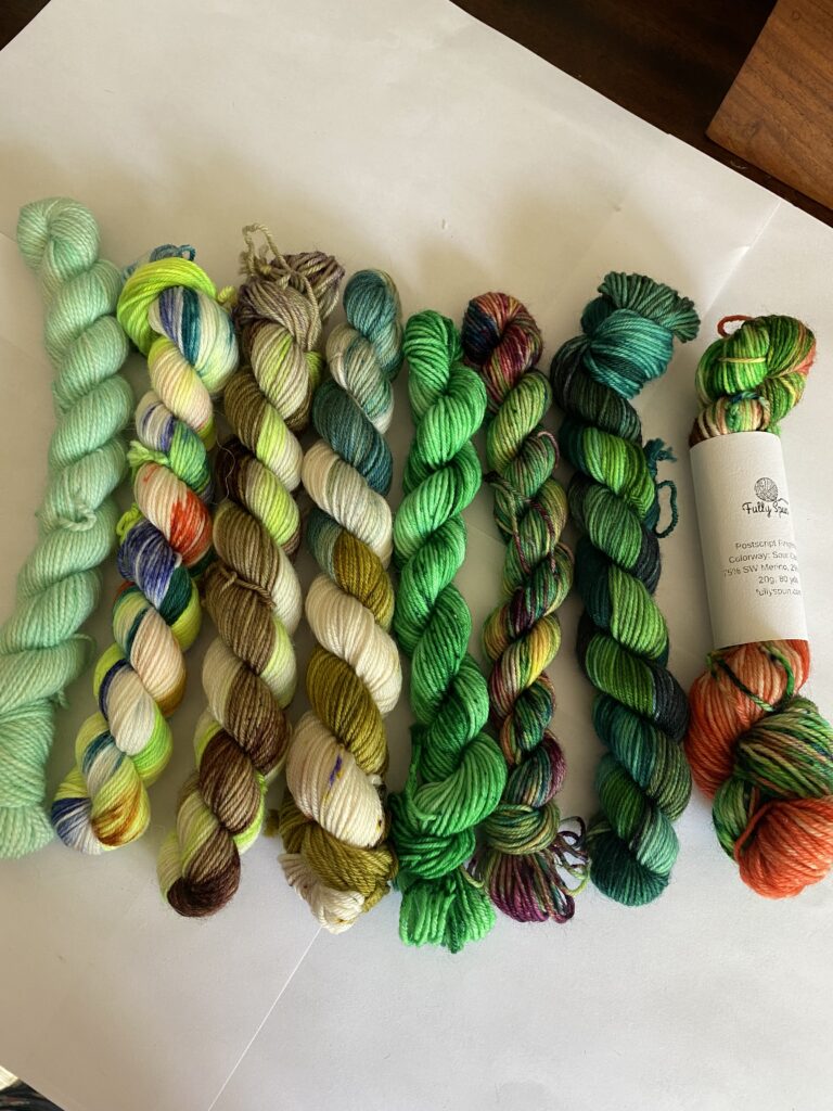 8 mini skeins All the Greens yarn with hints of orange, blue, brown and yellow.