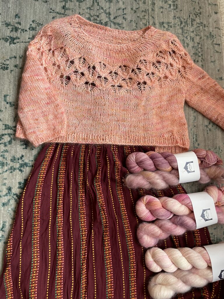 A flat lay of an outfit with a peach sweater, red and yellow striped dress and skeins of pink and purple yarn. 