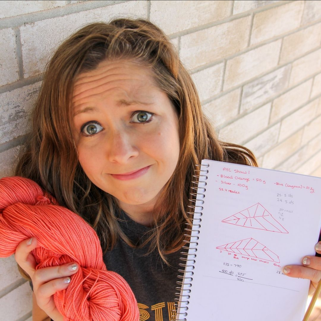 A white woman holding a notebook and hank of pink yarn looking a bit confused.